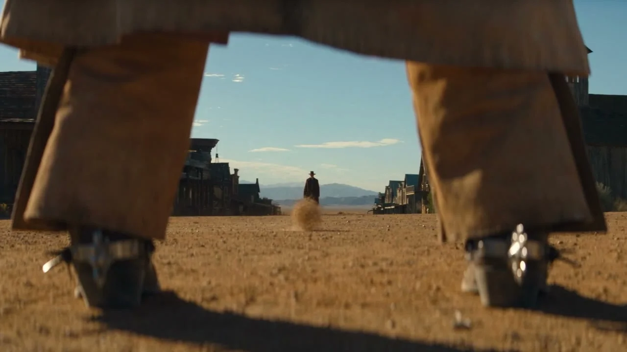 A dusty western street seen between a pair of cowboy boots, with a lone figure standing far down the road under a clear sky