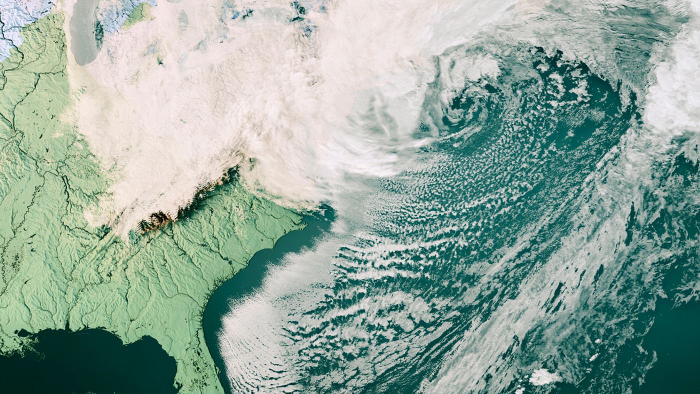 A satellite view of a storm swirling off the US east coast, with cloud over snowy land to the north and rows of small clouds streaming across the dark ocean