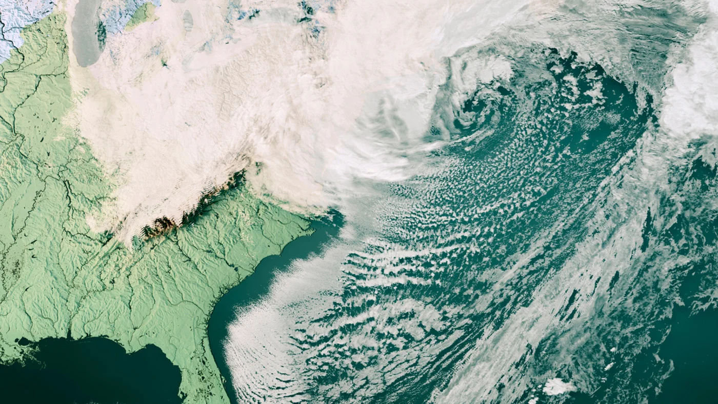A satellite view of a storm swirling off the US east coast, with cloud over snowy land to the north and rows of small clouds streaming across the dark ocean