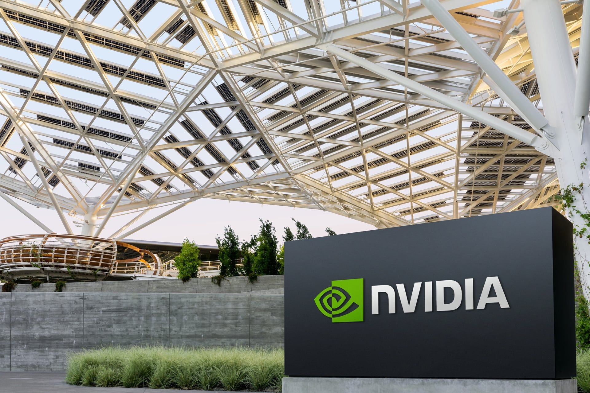The NVIDIA logo on a dark sign outside a building beneath a white lattice canopy of steel beams and solar panels
