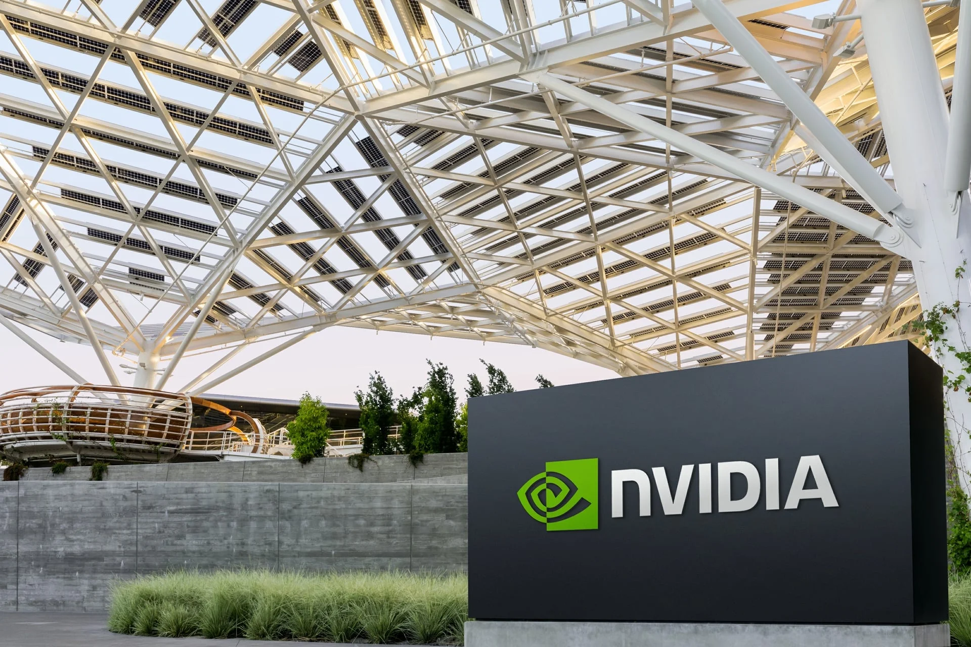 The NVIDIA logo on a dark sign outside a building beneath a white lattice canopy of steel beams and solar panels